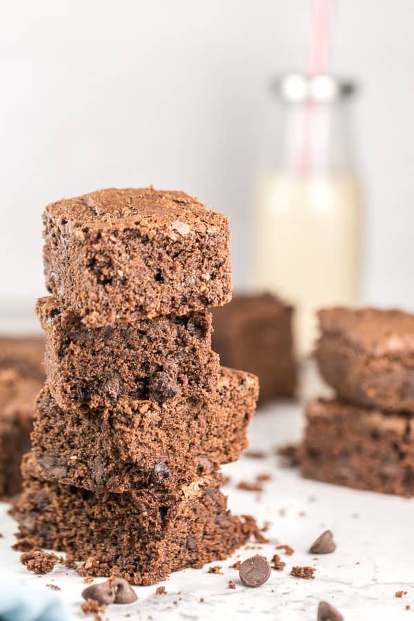 a stack of chewy brownies with additional brownies and a glass jug of milk in the background