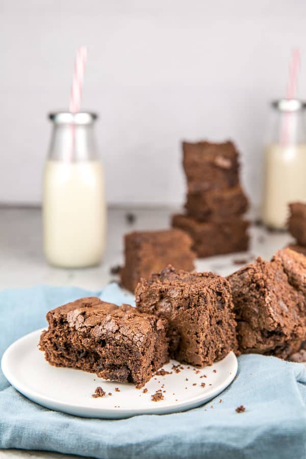a pile of rich brown chewy brownies on a white dessert plate with two glass bottles of milk in the background