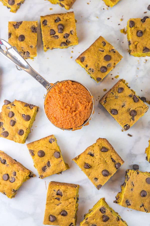 overhead view of pumpkin chocolate chip cookie bars cut into squares and scattered on a marble countertop surrounding a measuring cup filled with pumpkin puree.