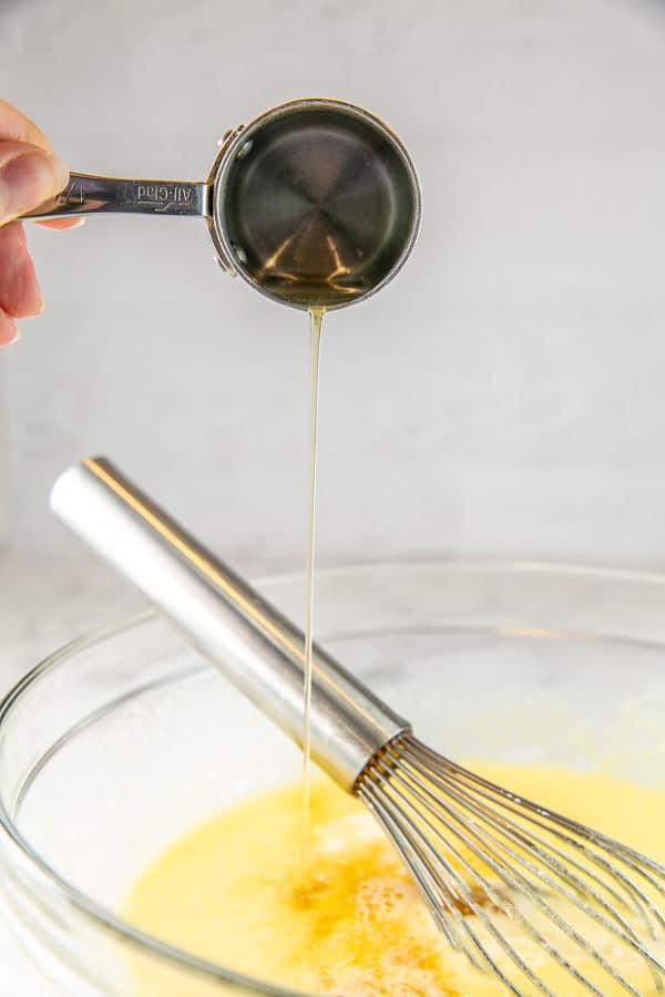 a stream of bourbon pouring from a small metal measuring cup into a glass mixing bowl full of buttermilk pie custard