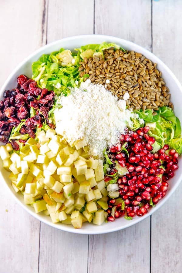 a large white salad bowl with shaved brussels sprouts and neat divided piles of dried cranberries, chopped apples, pomegranate arils, roasted sunflower seeds, and pecorino cheese.