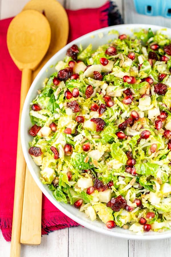 overhead view of a shaved brussels sprout salad in a white salad bowl with two bamboo salad spoons on a red cloth placemat.