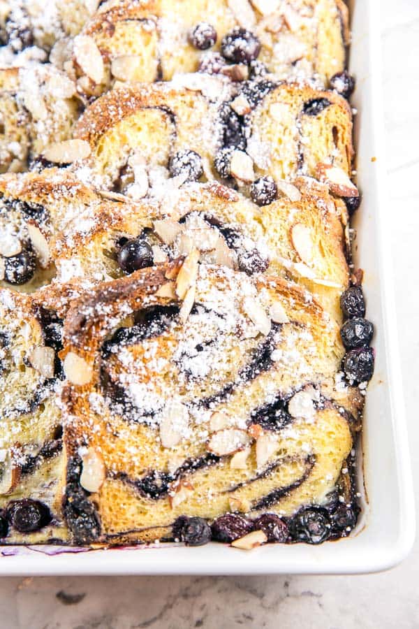 just baked babka french toast casserole covered in powdered sugar with blueberries and almond slices scattered on top.