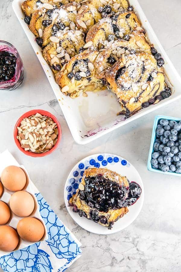 overhead view of a white casserole dish with baked babka french toast with one slice removed on a small plate covered in homemade blueberry sauce
