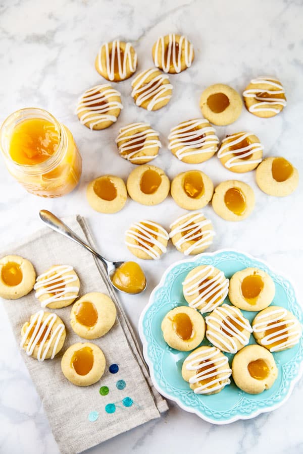 lemon thumbprint cookies covered in lemon glaze on a white marble counter with lemon curd and a blue dessert plate