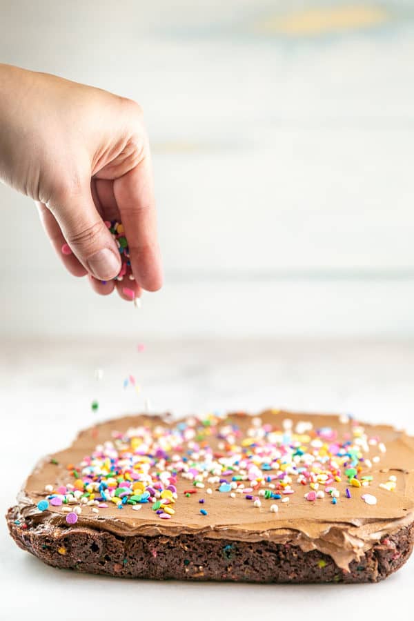 action shot of a hand spreading sprinkles on brownies covered with chocolate frosting