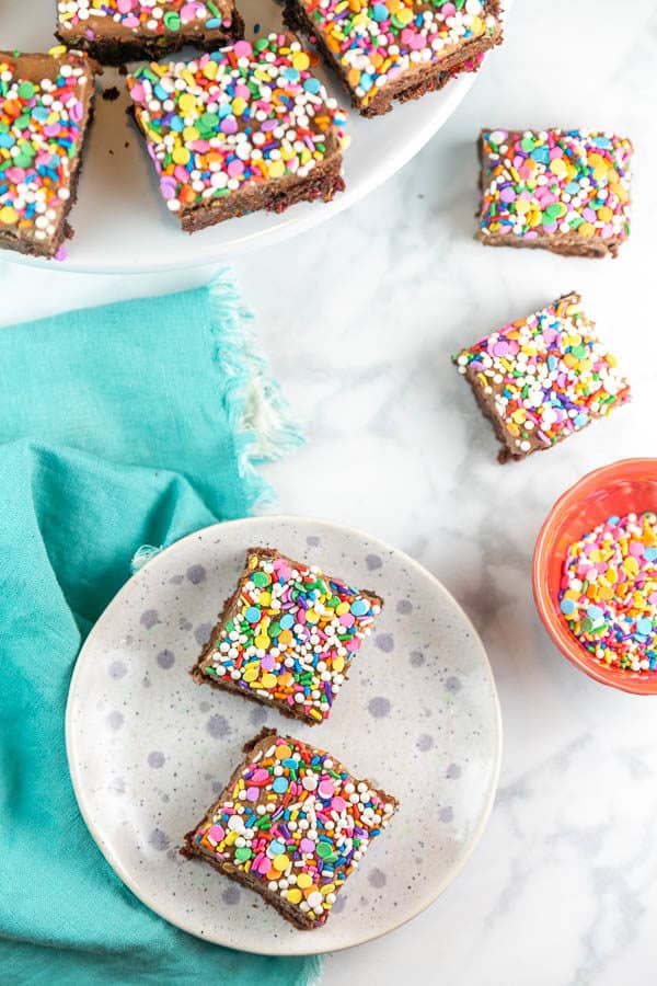 frosted sprinkle brownies on a small dessert plate, a white cake plate, and a marble counter, with extra sprinkles in a small decorate bowl