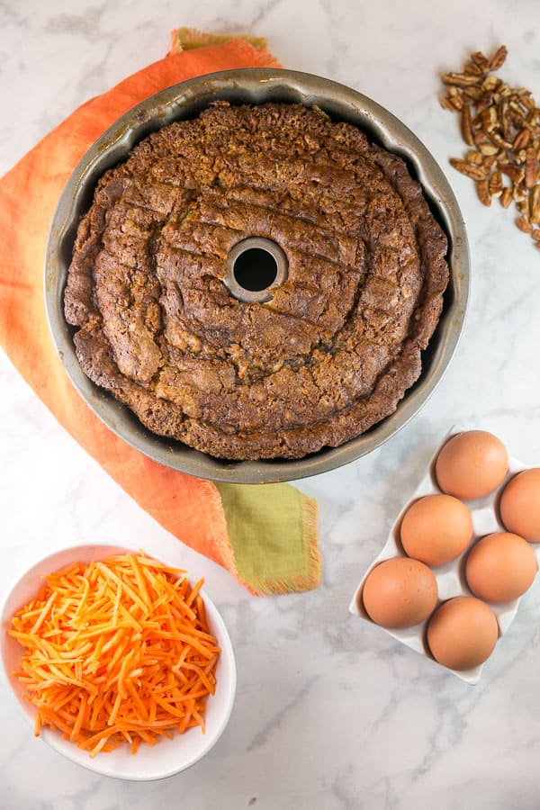 a baked carrot bund cake still in the bundt pan surrounded by carrots, eggs, and pecans.