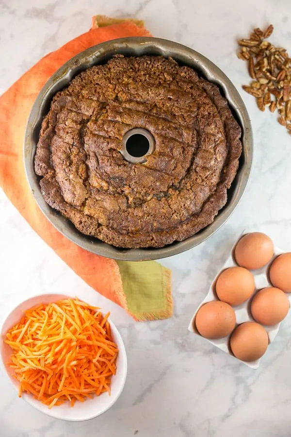 a baked carrot bund cake still in the bundt pan surrounded by carrots, eggs, and pecans.