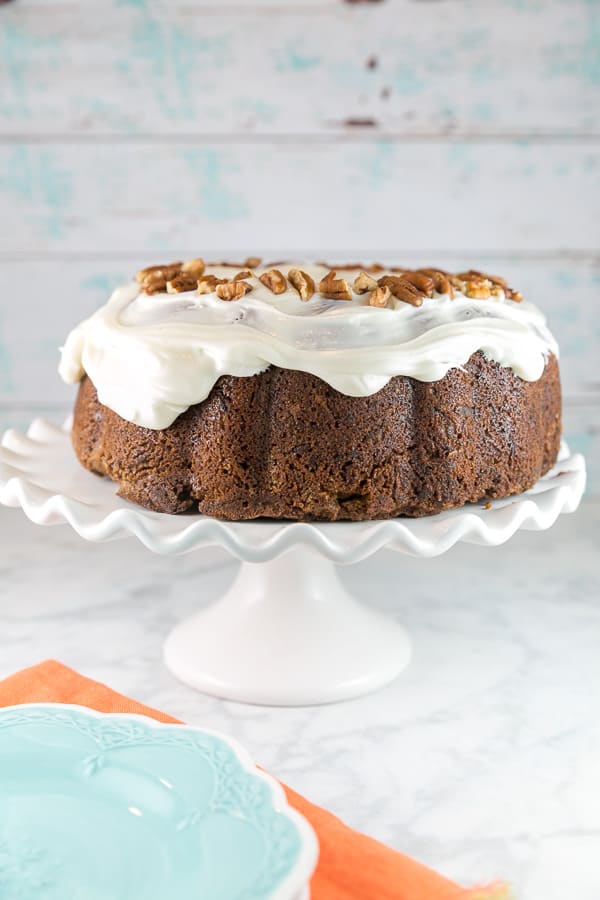 carrot bundt cake covered in cream cheese frosting on a white cake stand.