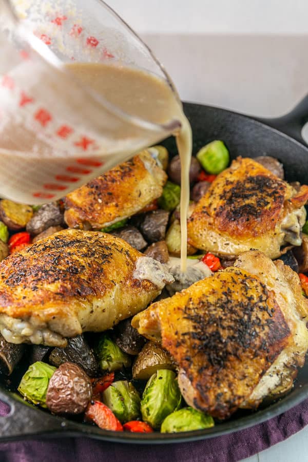 tahini sauce being poured into a cast iron skillet full of chicken, potatoes, and brussels sprouts