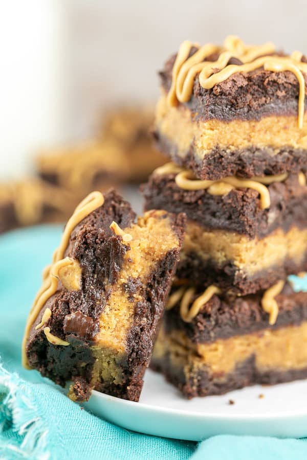 three peanut butter stuffed brownies with peanut butter drizzle stacked on a plate with another brownie with a bite taken out of it