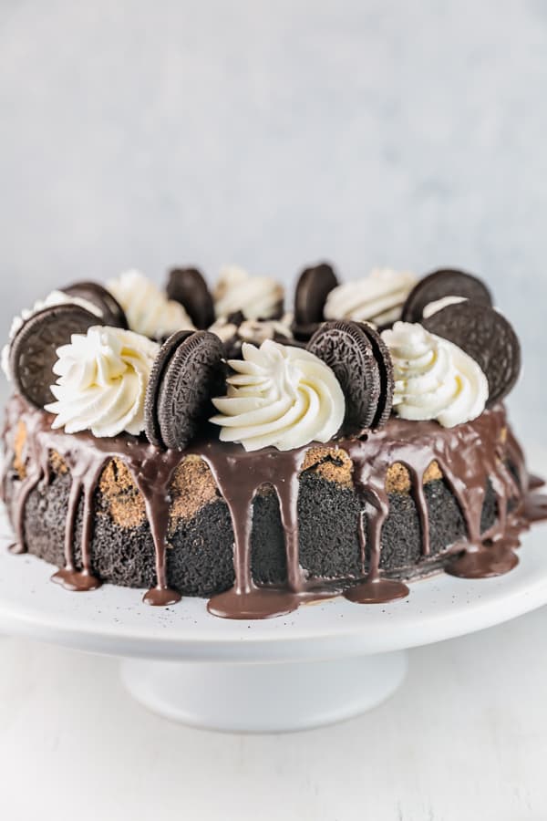an oreo cheesecake decorated with ganache, whipped cream, and oreos on a white cake stand