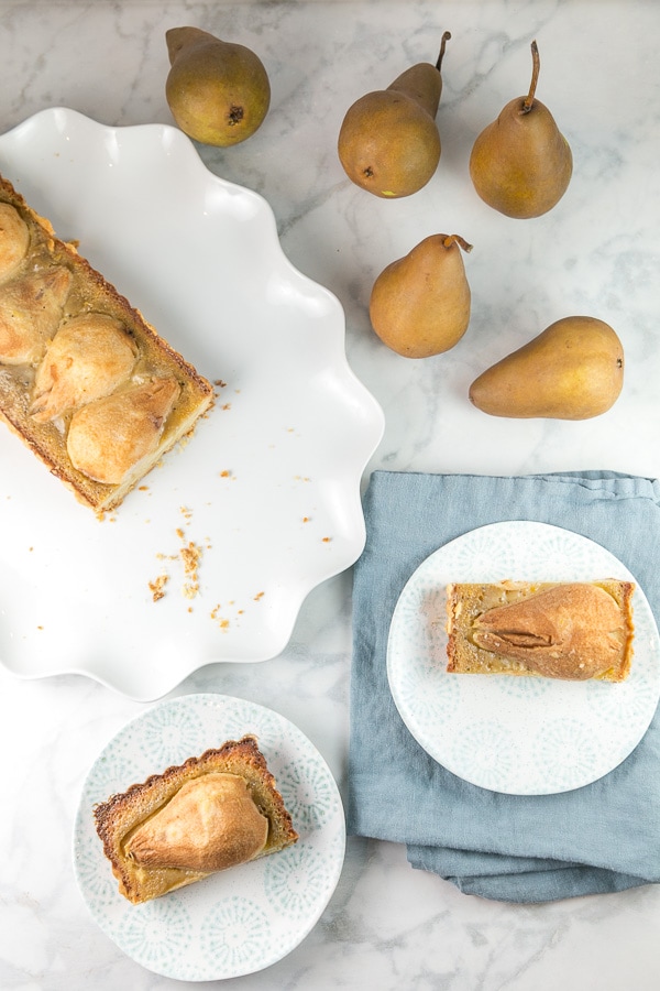 two pieces of pear tart on dessert plates surrounded by fresh pears.