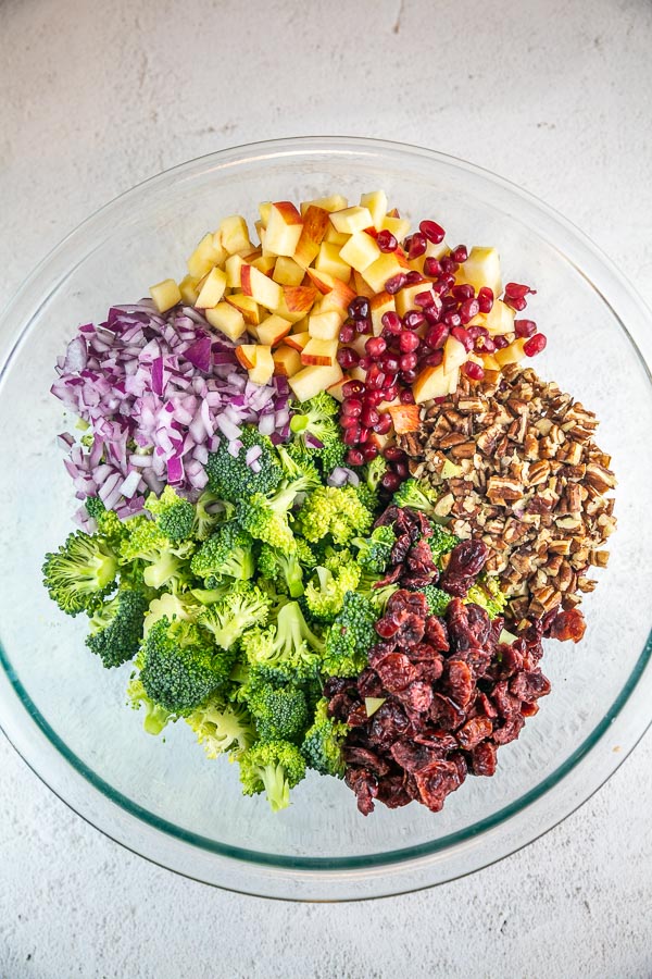 broccoli florets, red onion, apples, pomegranate arils, pecans, and cranberries in a glass bowl