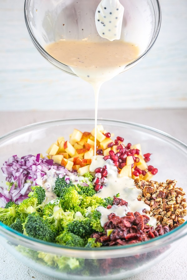 dressing being poured from a mixing bowl onto broccoli salad ingredients