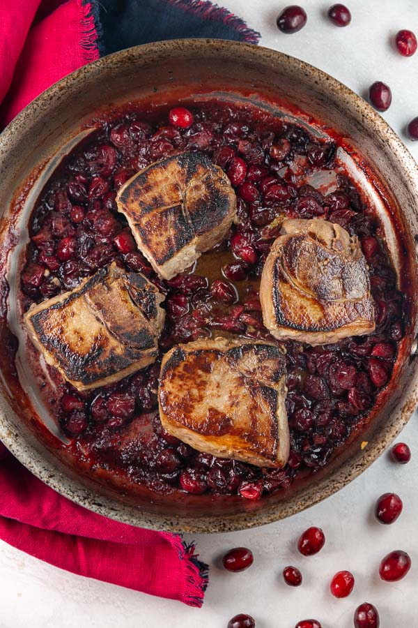 four seared pork chops in a pan with a cranberry maple sauce.