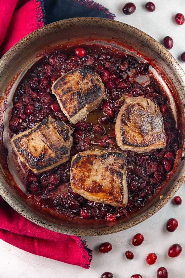 four seared pork chops in a pan with a cranberry maple sauce.