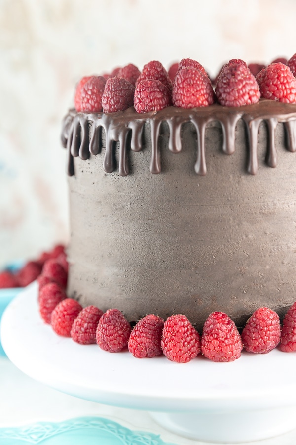 three layer chocolate cake with chocolage ganache drips surrounded by fresh raspberries