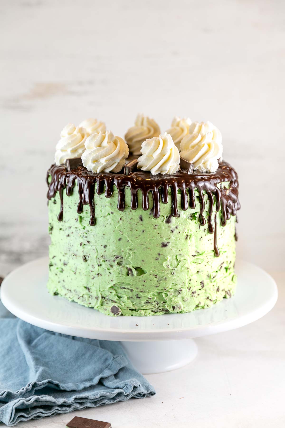 three layer cake covered in green mint chocolate chip frosting with chocolate ganache drips and whipped cream swirls