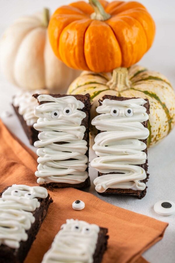 two brownies covered with white chocolate drizzle and candy eyeballs leaning against small pumpkins