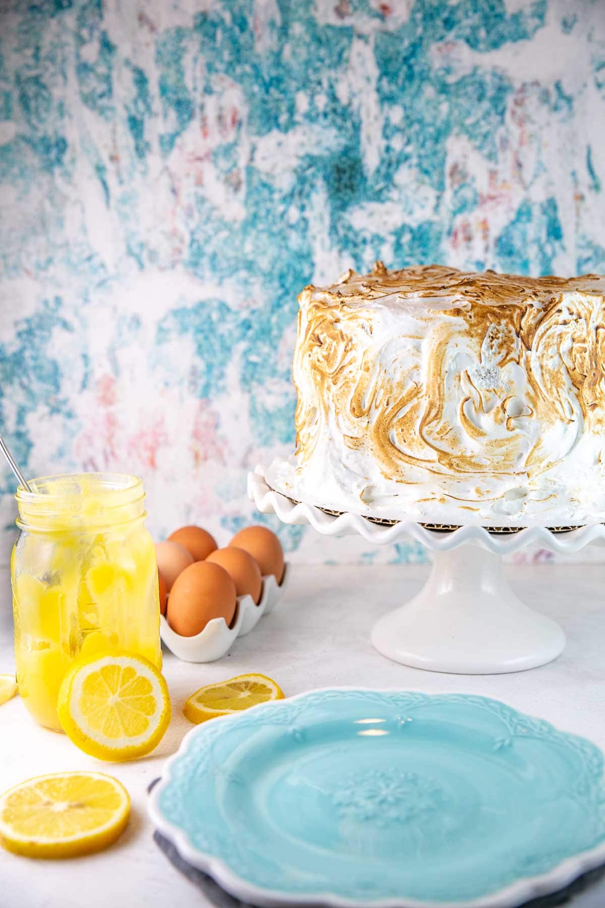 three layer cake covered with toasted meringue with eggs and lemon curd in the background
