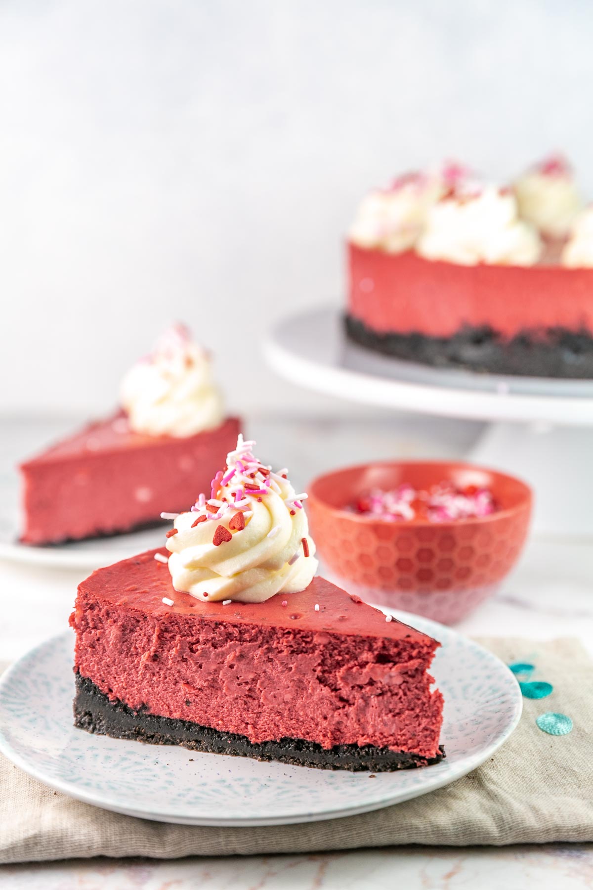 slice of red velvet cheesecake on a dessert plate with an additional slice and sprinkles in the background