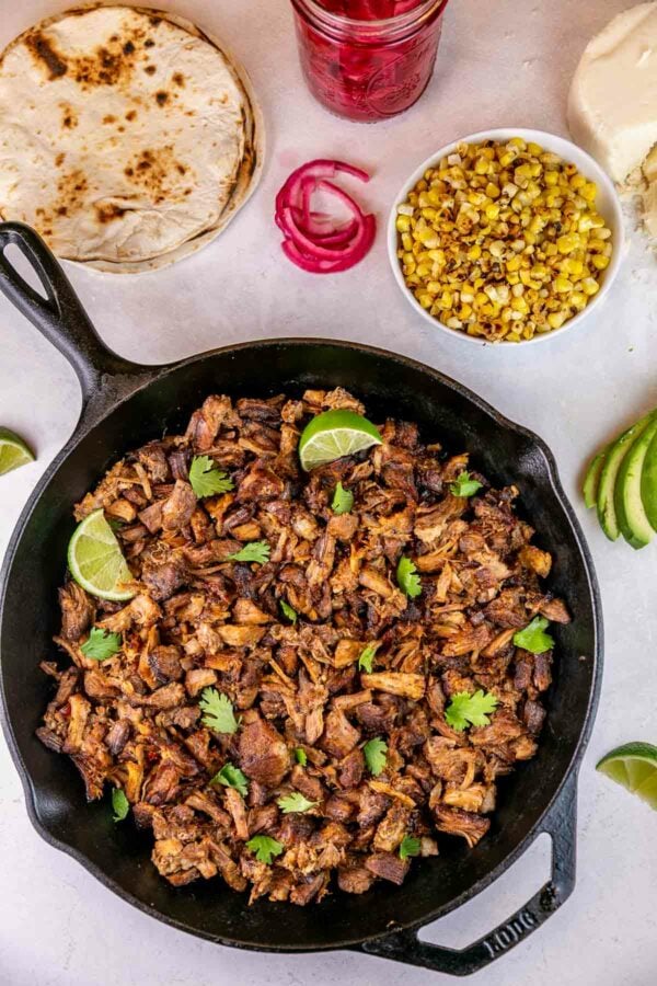 carnitas in a skillet surrounded by tortillas, charred corn, red onion, and avocado
