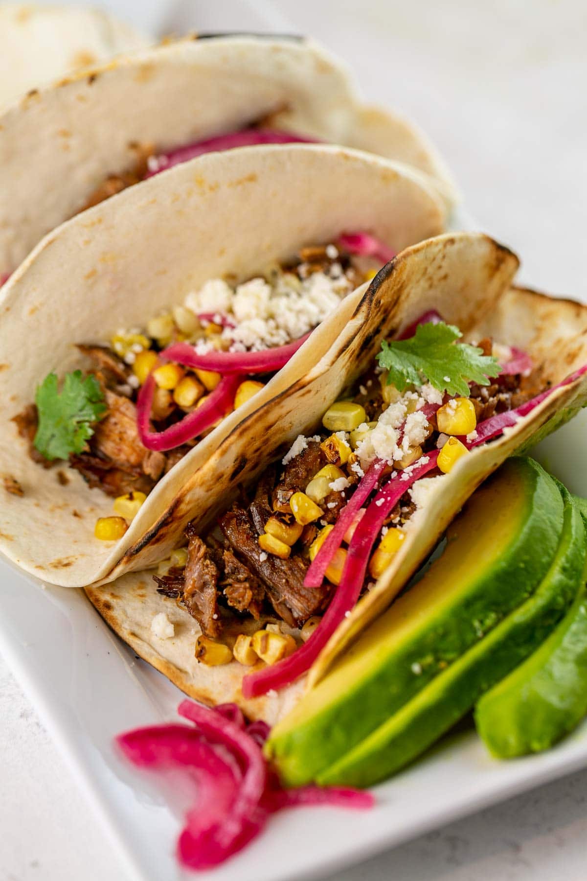 carnitas in soft flour tortillas topped with red onion, corn, and cojita