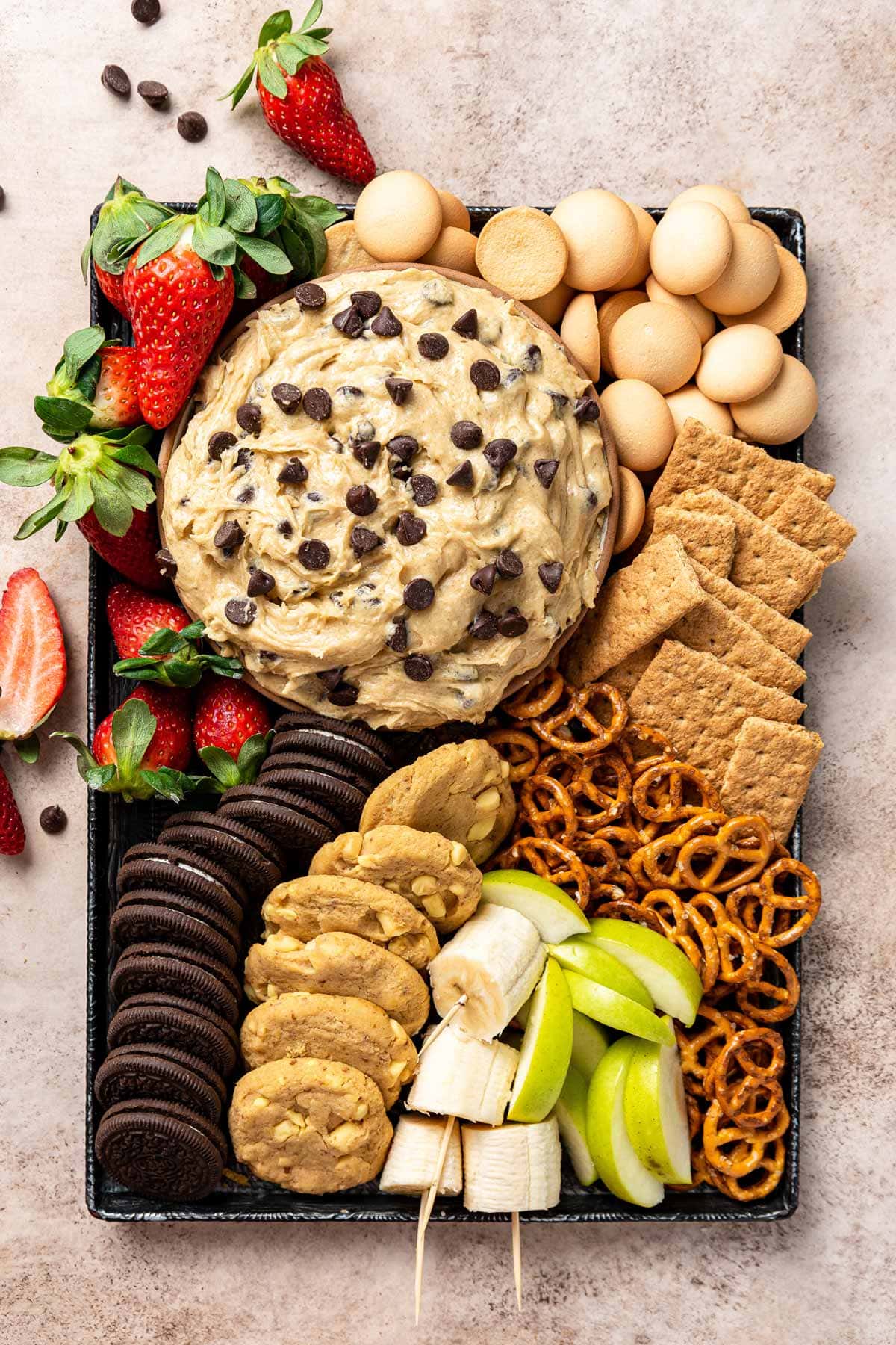 bowl of cookie dough dip surrounded by cookies, pretzels, and fruit