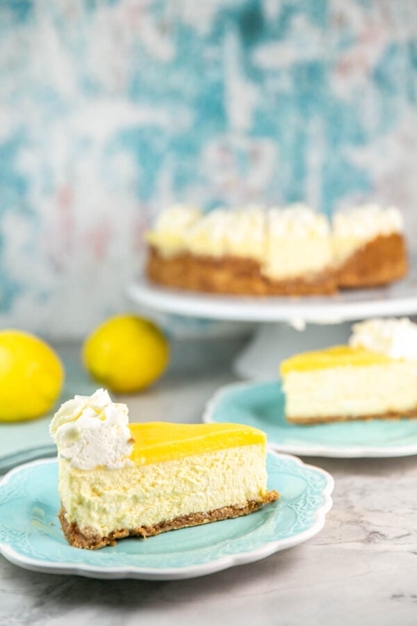 Slice of lemon cheesecake with the entire cheesecake in background.
