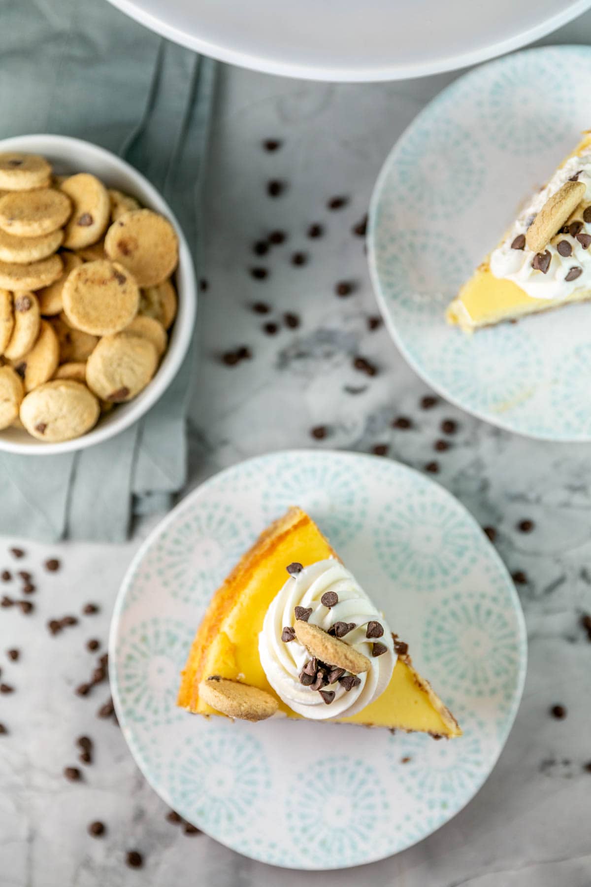 Top view of slide of cheesecake on a white plate topped with whipped cream, chocolate chips and a cookie. 