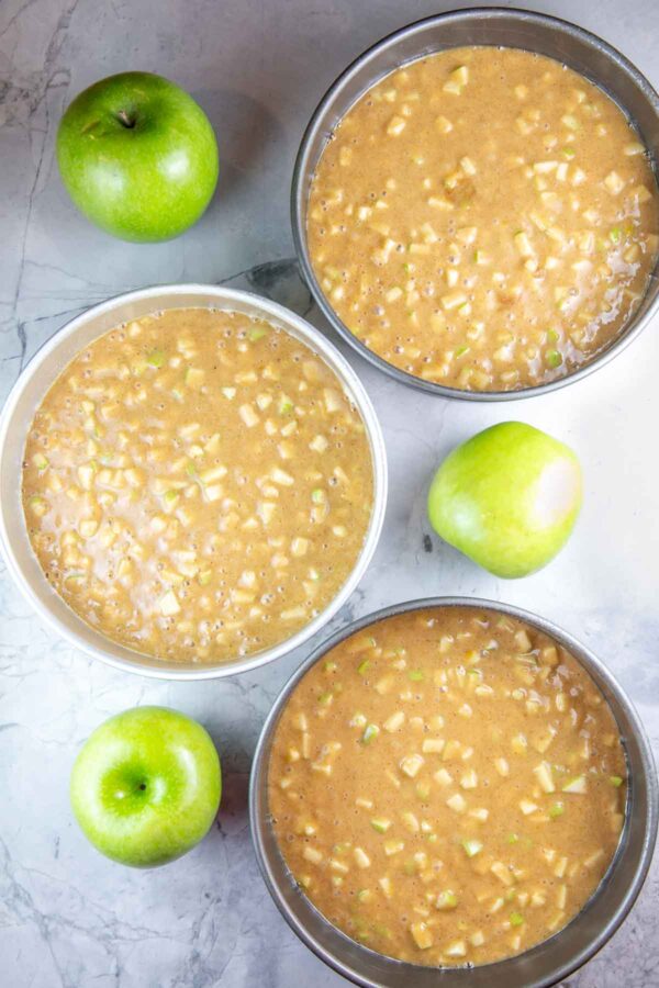 Three cake pans filled with apple cake batter sitting next to whole apples. 