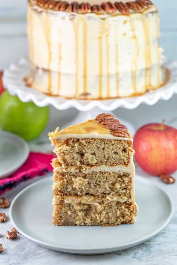 Slice of cake on a grey plate with apples and full cake in the background. 