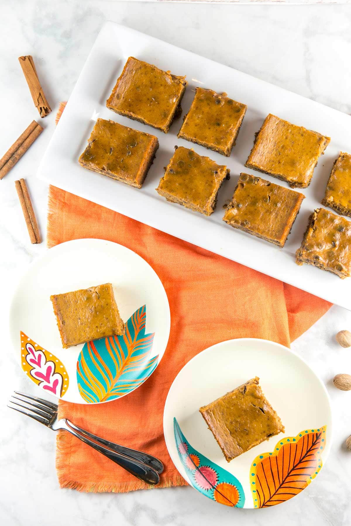 overhead  view of pumpkin pie bars on serving plates surrounded by whole nutmeg and cinnamon sticks.