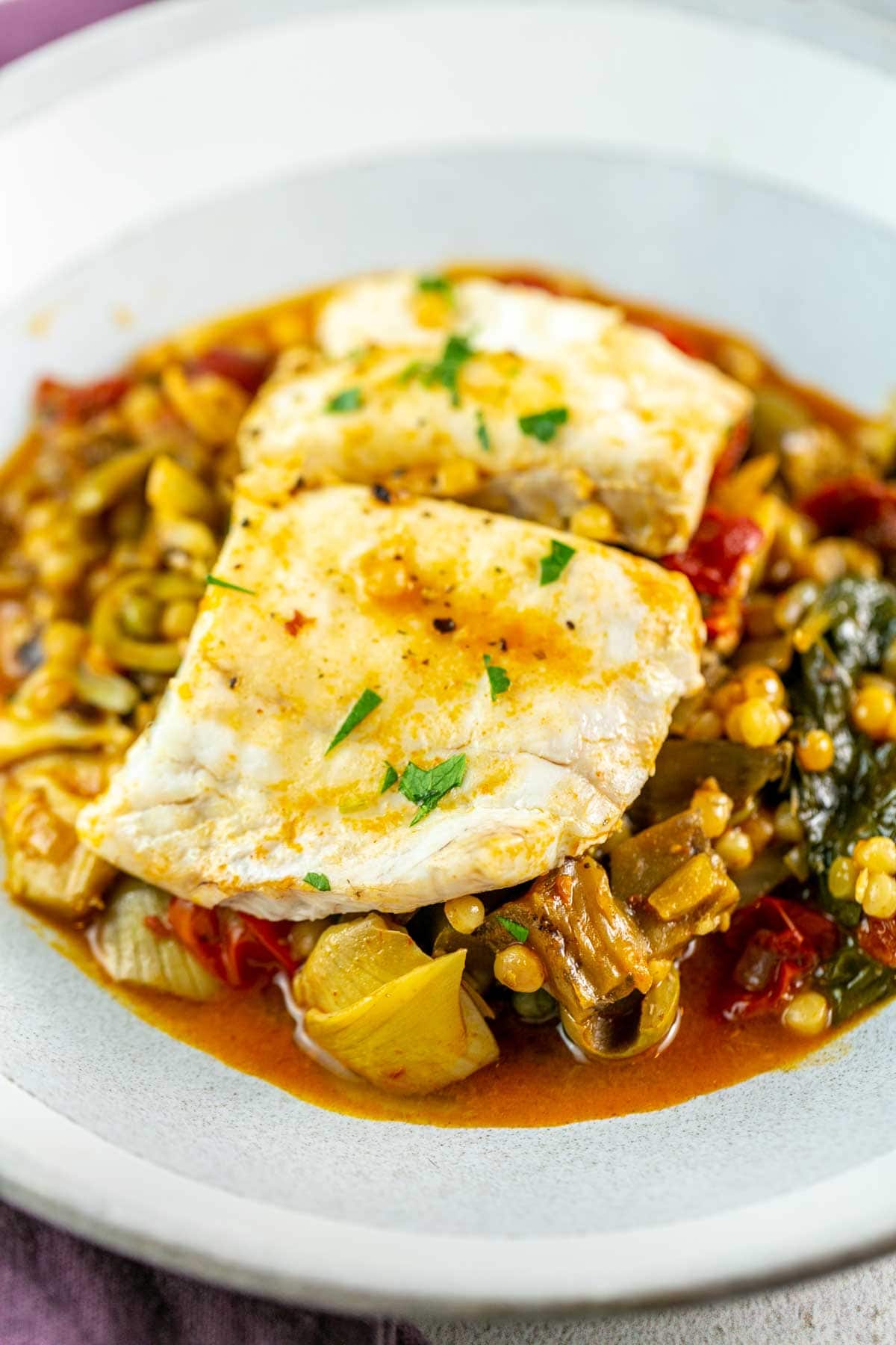 Mediterranean poached fish on bed of couscous and vegetables on a grey plate.