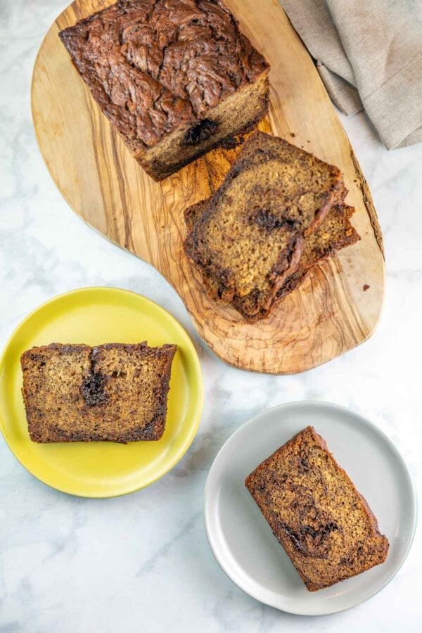 Top view of two plates with slices of banana bread on them and the rest of the loaf on a wooden cutting board. 
