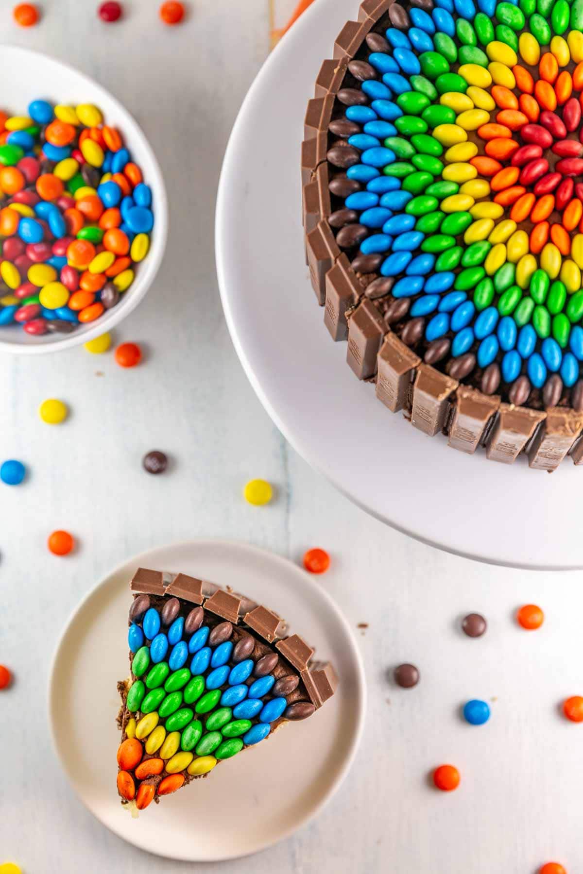Top view of slice of candy cake with the whole candy cake next to it and a bowl of m&ms.