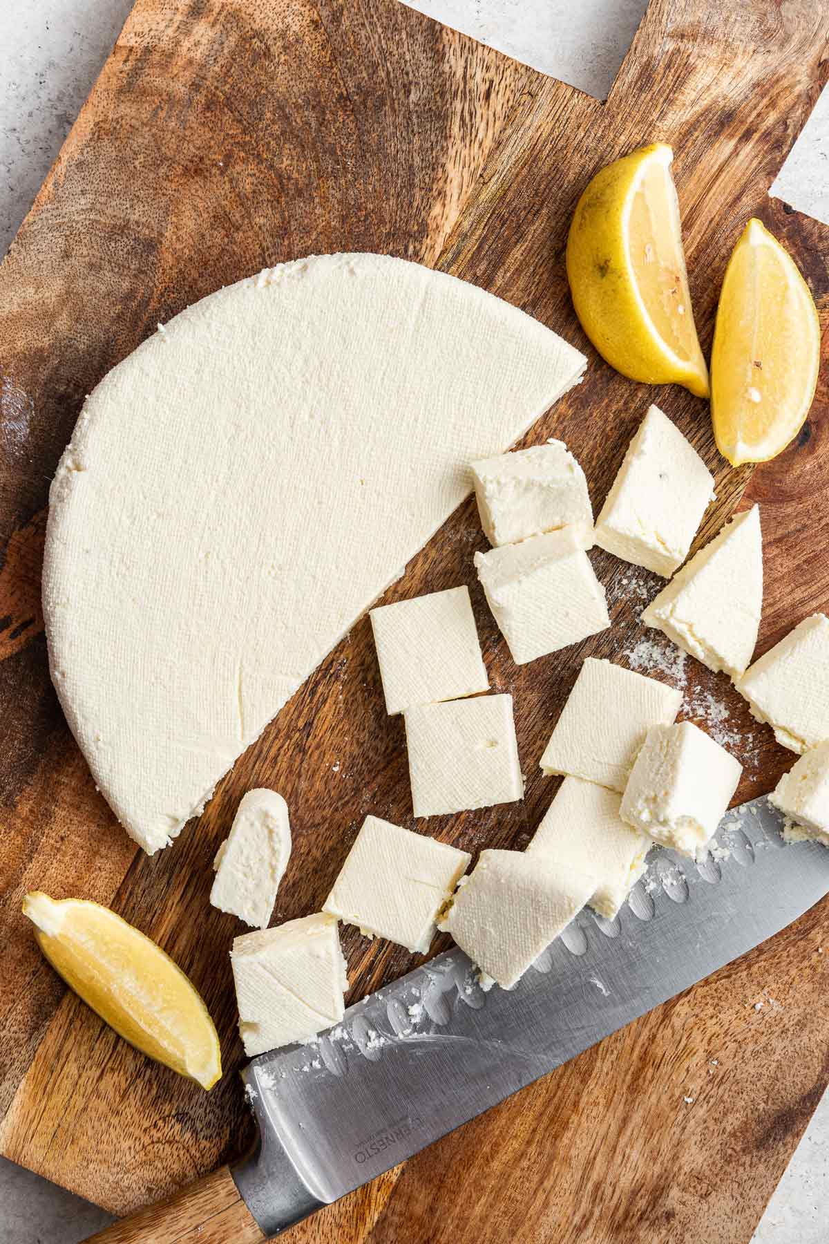 Paneer block of cheese with half of the cheese cut into smaller pieces on a wooden cutting board. 