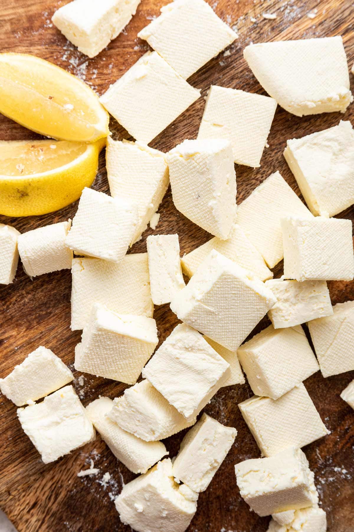 Small pieces of paneer on a wood cutting board next to lemon wedges. 