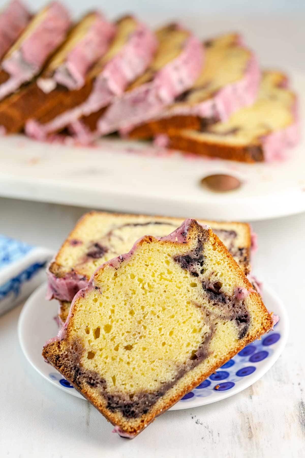 Slice of blueberry lemon pound cake on a white plate with a stack of more slices behind it. 