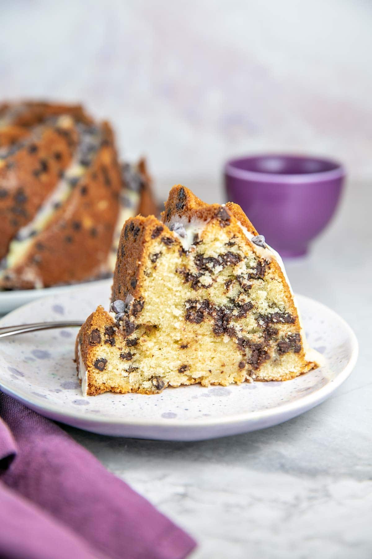 Slice of chocolate chip bundt cake on a white speckled plate with whole cake in background.