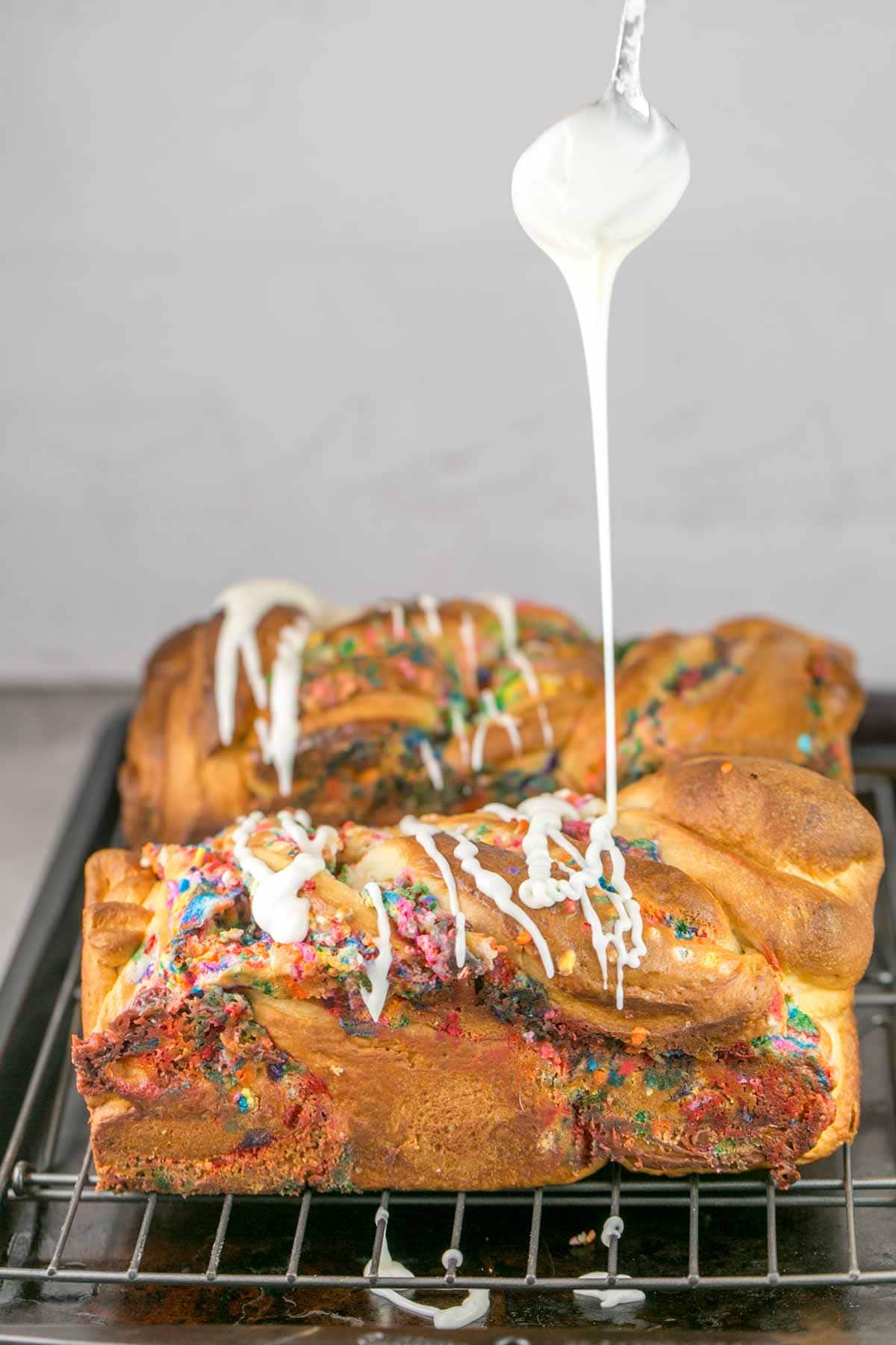 smooth vanilla glaze dripping from a spoon down onto two loaves of funfetti babka