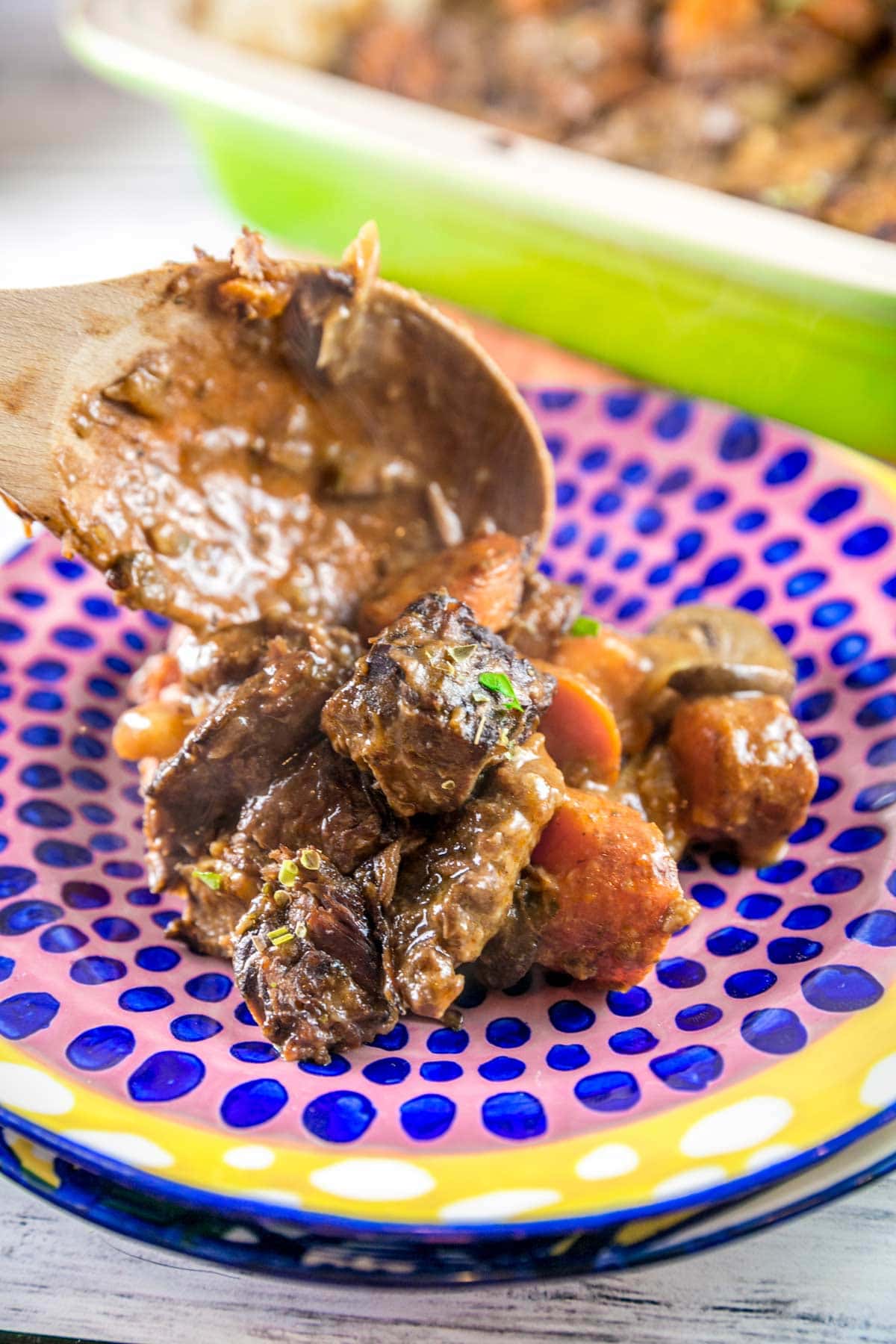 a wooden spoon dishing beef stew onto a brightly colored polka dotted pasta bowl