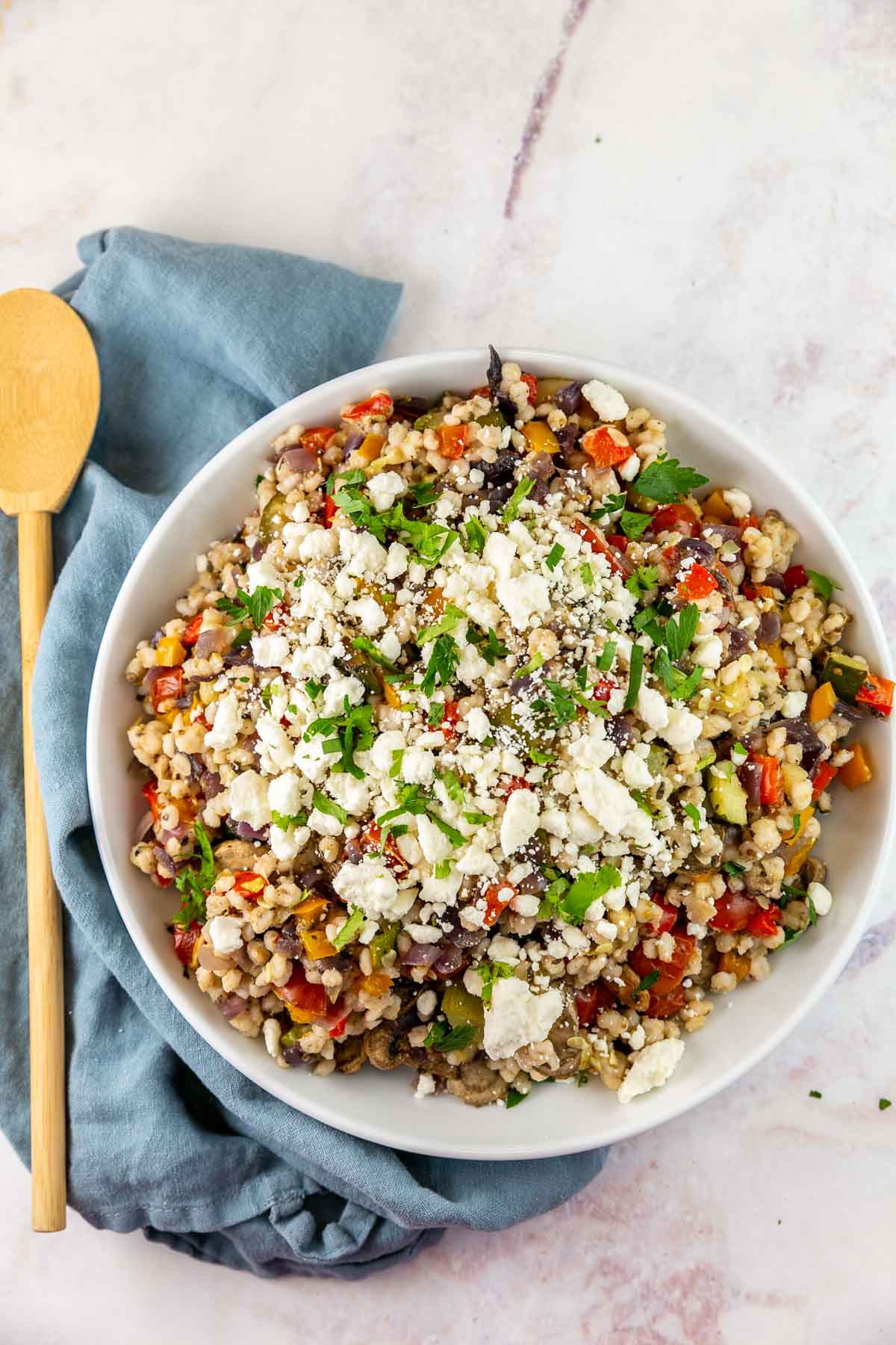 Top view of Mediterranean barley salad topped with feta cheese in a white bowl with a wooden spoon on the side. 