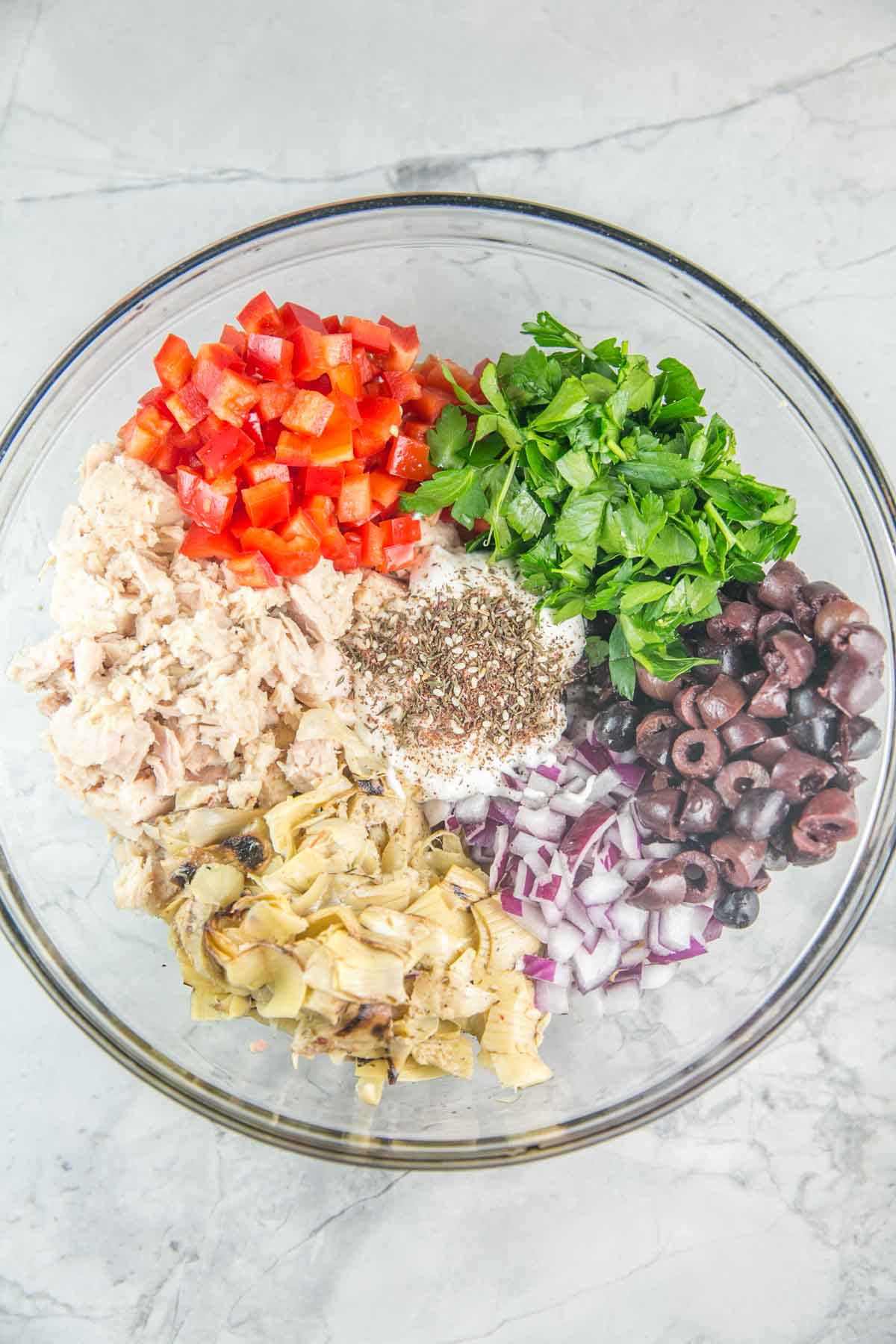 overhead view of a glass mixing bowl with piles of light chunk tuna, red bell pepper, artichoke hearts, red onion, greek olives, parsley, mayo, and italian herbs