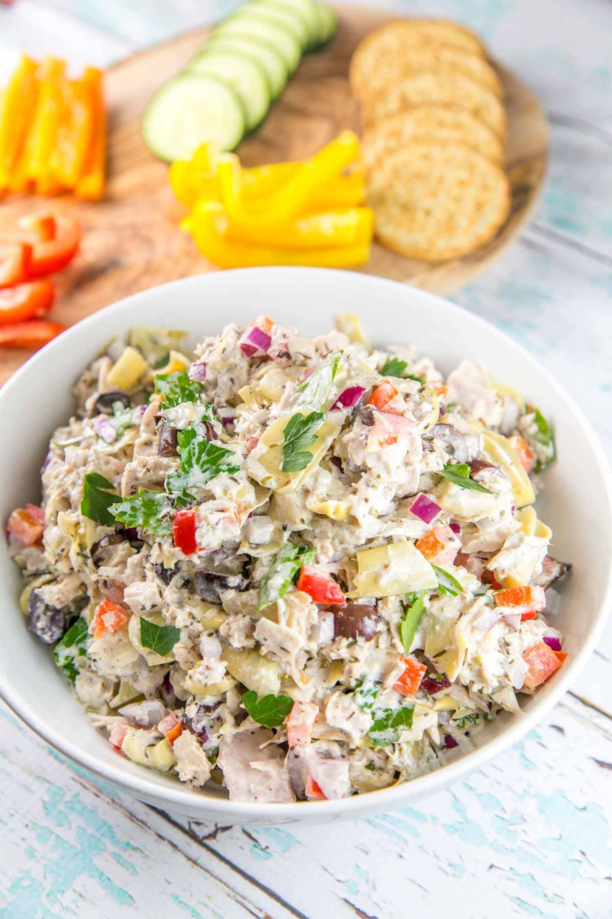 a large white bowl full of mediterranean tuna salad with cut vegetables and crackers in the background