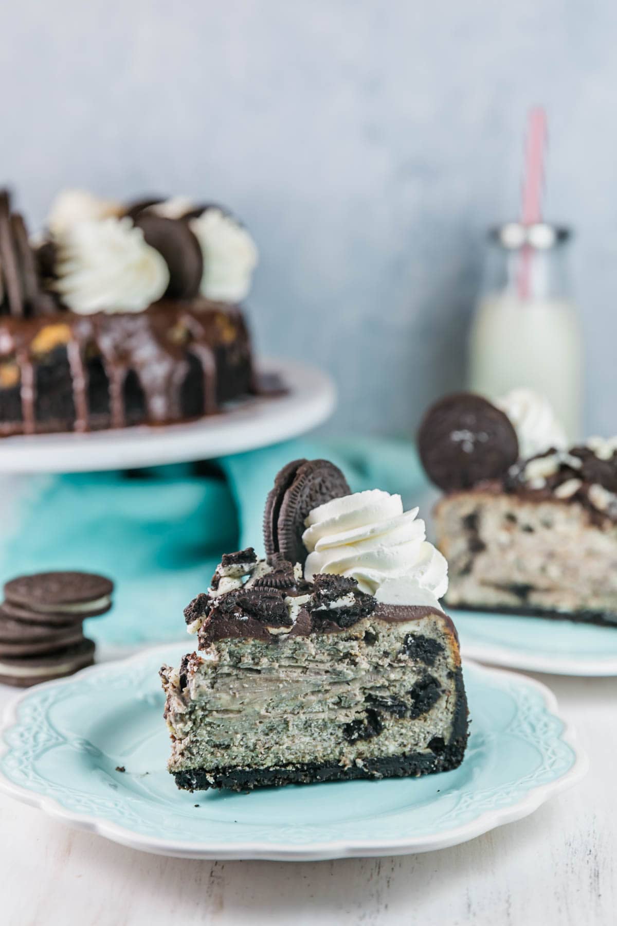 two slices of oreo cheesecake on dessert plates with oreos and a glass milk jug in the background