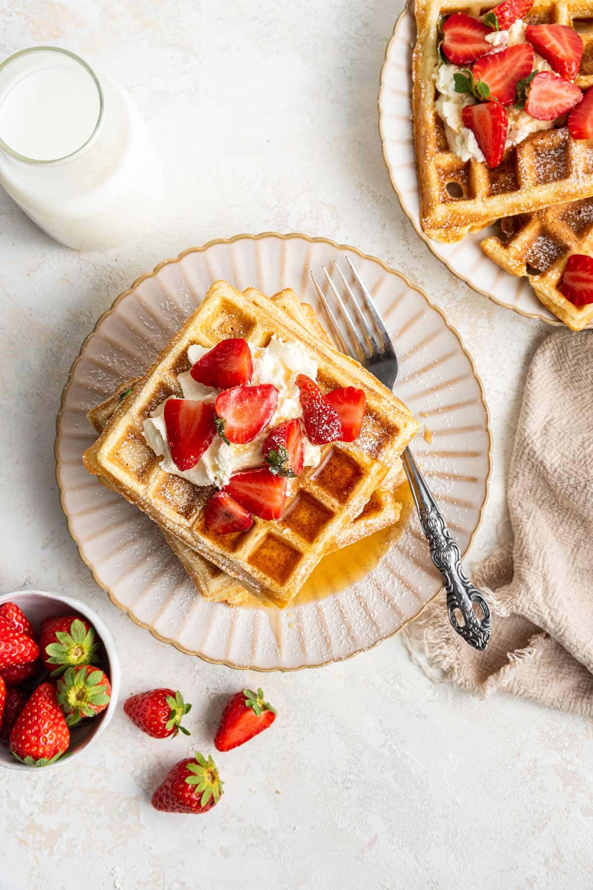 a stack of waffles covered with whipped cream, strawberries, maple syrup, and a dusting of powdered sugar