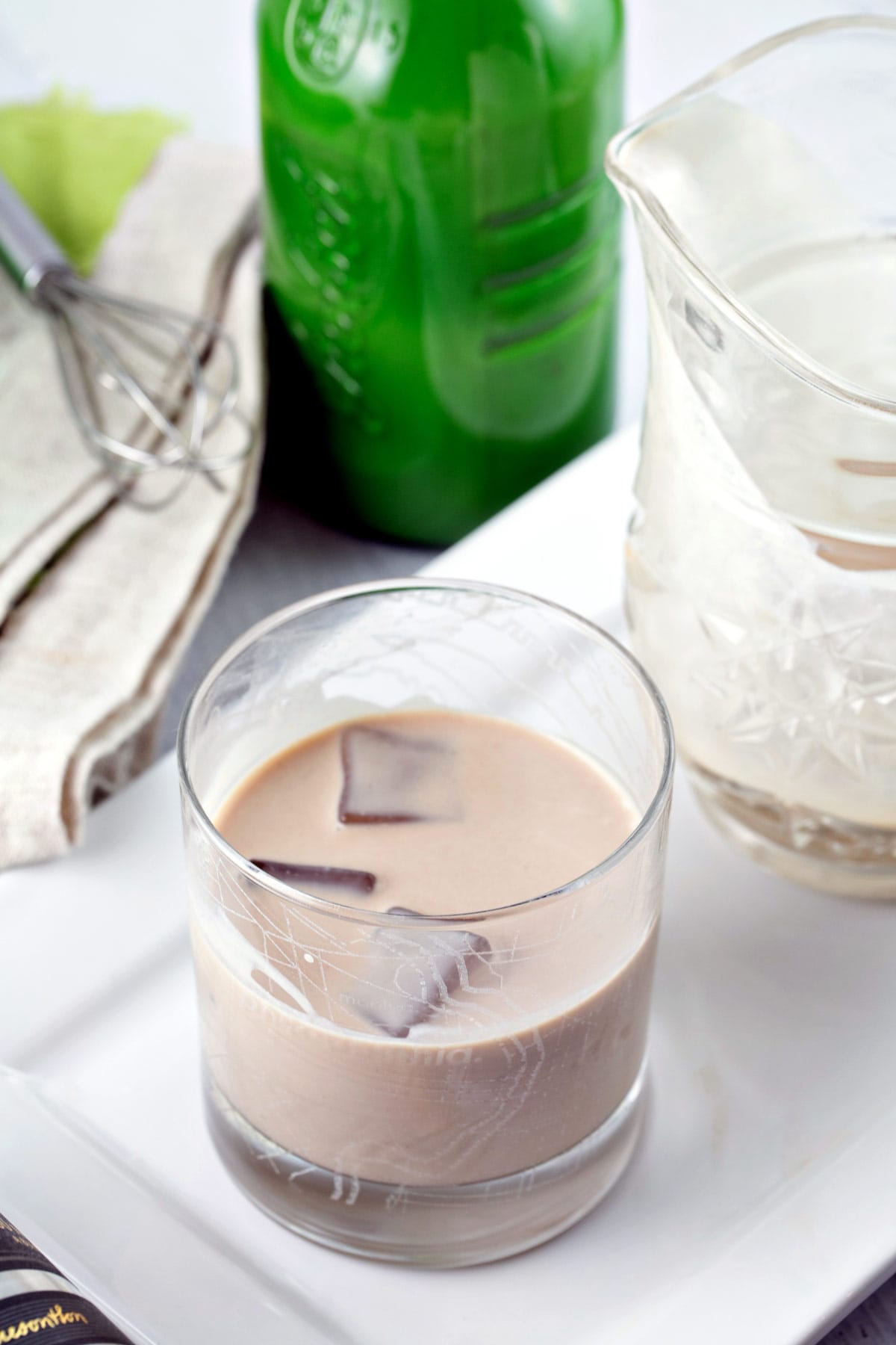 lowball glass with ice cubes and homemade irish cream in front of a green glass bottle